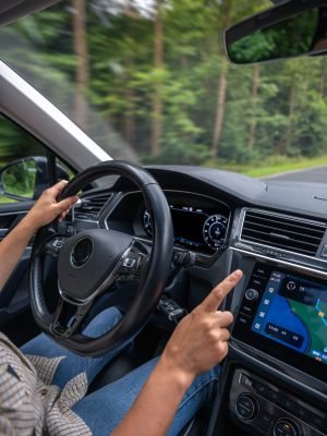 Vertical photo of a female driver interacting with the touchscreen navigation system while driving through a forest road with blurred trees.